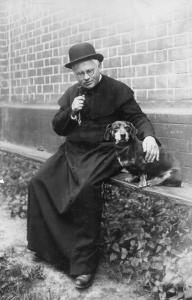 Juozas Tumas-Vaižgantas su šuniuku Kauku prie bažnyčios, 1928 m. Nežinomas XX a. I p. fotografas. Maironio lietuvių literatūros muziejaus nuosavybė